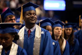A young man in a blue graduation cap and gown with a white stole and a University of Kentucky logo stands among fellow graduates at a commencement ceremony. He gazes upward with a proud and reflective expression. Other graduates in blue caps and gowns are visible in the background, slightly out of focus, in a large indoor arena with bright lighting.